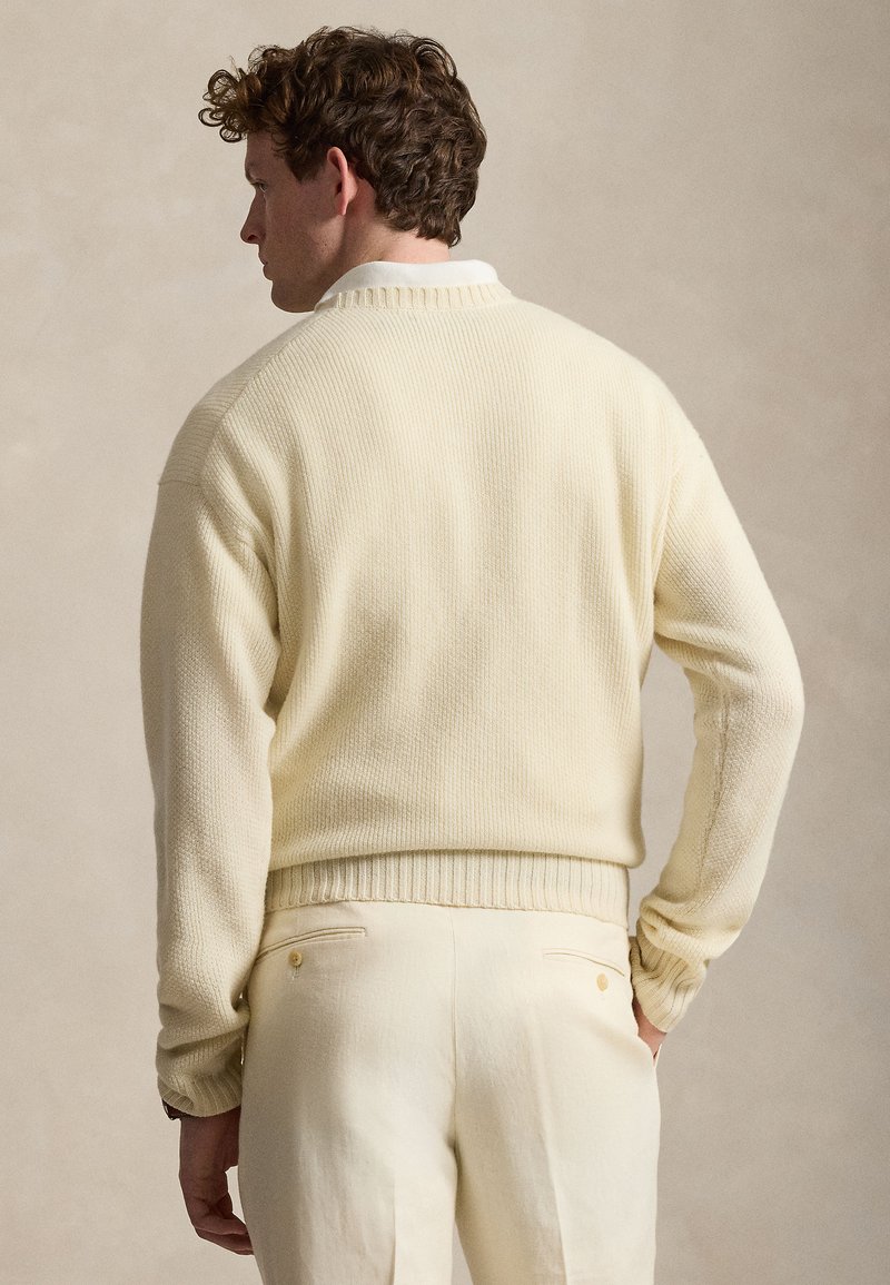 Uomo con capelli ricci visto di spalle, indossa un maglione lavorato a maglia color crema sopra una camicia bianca con colletto e pantaloni color crema.