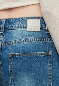 Blå denimbukser med synlig syning, et læderlignende mærke på taljen med teksten "OBJECT" og to baglommer.