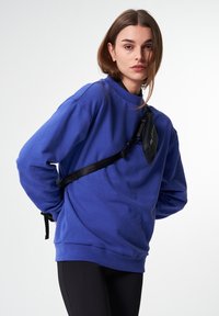 Blaues Sweatshirt mit lockerer Passform, langen Ärmeln und Rundhalsausschnitt. Ein schwarzer Riemen mit einer kleinen Tasche kreuzt sich über die Vorderseite.