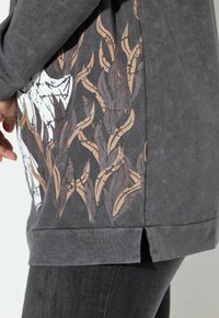 Maglietta grigia a maniche lunghe con tessuto testurizzato, caratterizzata da un design laterale con linee intrecciate marroni e beidge, e una grafica bianca sul davanti.