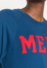 Blå bomullströja med en röd "M"-grafik. Har rund halsringning, raglanärmar och texturerad stickad detalj.
