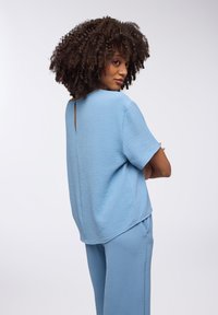 Top azzurro chiaro testurizzato con maniche corte e dettaglio a oblò sul retro, abbinato a pantaloni leggeri coordinati. Design semplice e rilassato.