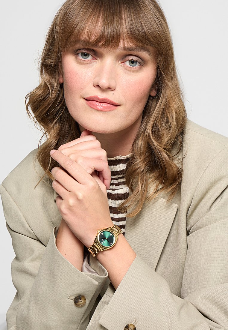 Montre bracelet en or avec un cadran rond vert, un bracelet en métal et une finition brillante. La personne porte un blazer beige et un haut rayé.