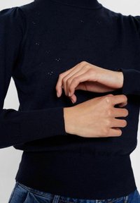 Persoon die de manchet van een marineblauwe trui met kleine gaatjes aanpast, gekleed in blauwe jeans, tegen een effen lichte achtergrond.