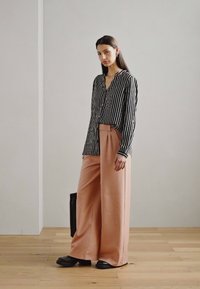 Black and white striped long-sleeve shirt paired with wide-legged pink trousers. Model holds a black bag. Floor is wooden, background is plain.