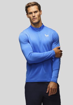 Homme aux cheveux courts portant un haut de sport bleu à manches longues avec une fermeture éclair quart et un logo blanc sur un fond gris clair.