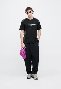 Μαύρο μπλουζάκι Nike Shox με γκρι λεπτομέρειες, μαύρη φόρμα και γκρι αθλητικά παπούτσια. Κρατάει ένα ροζ καπέλο. Μοντέλο με σγουρά μαλλιά και γυαλιά ηλίου.