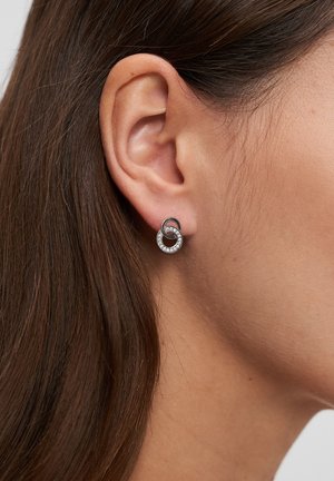 Boucles d'oreilles en argent en forme de créoles avec un design entrelacé, ponctuées de cristaux clairs le long du bord extérieur, sur un fond neutre.