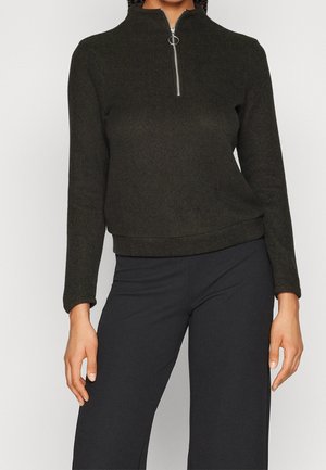 Femme portant un pull en maille foncé à manches longues avec une fermeture éclair argentée et un anneau, associé à un pantalon large et foncé.