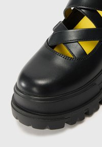 Black leather shoe with a rounded toe, cross-strap design, and a thick textured sole. Interior features yellow accents.