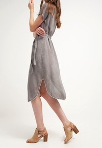 Harmaa mokkanahkainen midi-mekko lyhyillä hihoilla, vyötärösoljella ja epäsymmetrisellä helmalinjalla. Yhdistettynä ruskeisiin korkokenkiin.