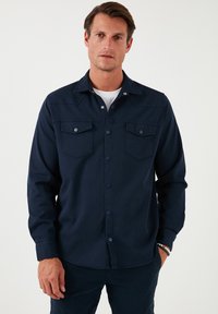 Cămașă bleumarin cu nasturi, fabricată dintr-un material neted. Are două buzunare pe piept cu clapete și guler. Mâneci lungi, design minimalist.