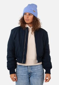 Giubbotto bomber navy con polsini a coste, tasche laterali e dettagli con cerniera sopra un hoodie beige chiaro. Indossato con un berretto lavorato a maglia bleu chiaro.