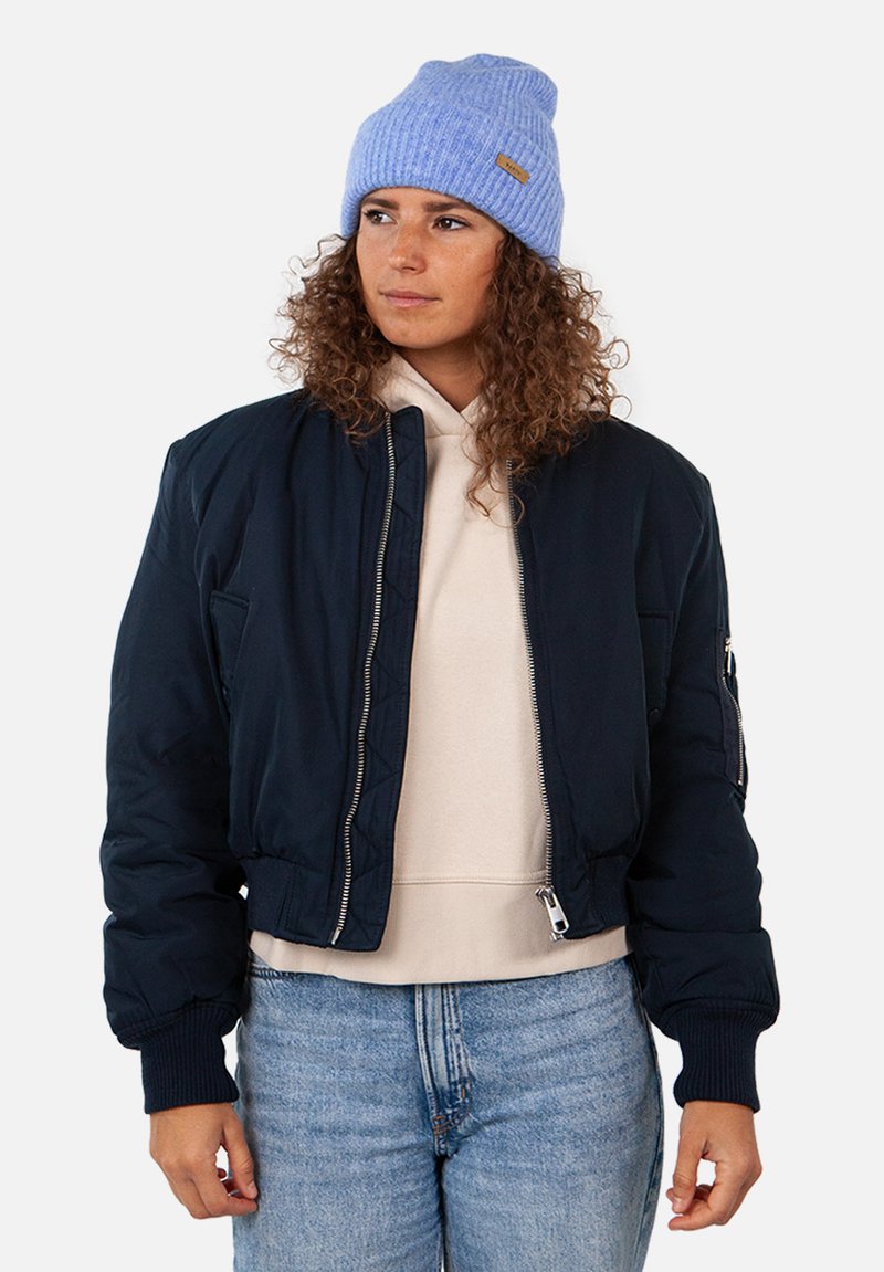 Giubbotto bomber navy con polsini a coste, tasche laterali e dettagli con cerniera sopra un hoodie beige chiaro. Indossato con un berretto lavorato a maglia bleu chiaro.