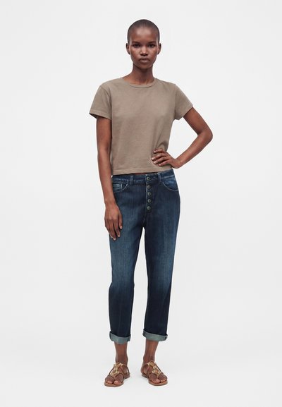 Femme debout, main sur la hanche, portant un t-shirt marron à manches courtes, un jean foncé retroussé avec des boutons et des sandales marron sur fond blanc.
