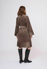 Nő barna kordbársony övvel átkötött kabátban és fekete térdig érő csizmában, háttal áll egy egyszerű világos háttér előtt.