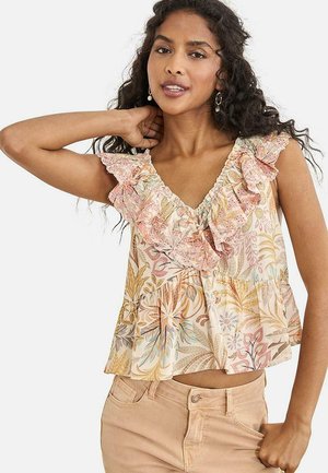 Mujer con cabello largo y rizado, vistiendo un top sin mangas con estampado floral y volantes de encaje, y pantalones beige, posando con una mano detrás de la cabeza.