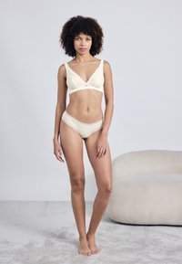 Σετ λευκής δαντέλας σε κρέμα χρώμα με ραφή χωρίς μπράτσο και αντίστοιχα σλιπ χιπστερ, τονίζοντας τον μαλακό ύφασμα και την αόρατη σχεδίαση.