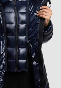 Chaqueta acolchada en color navy con un acabado brillante, diseño acolchado, dos cremalleras frontales y un ribete negro contrastante a lo largo de los lados.
