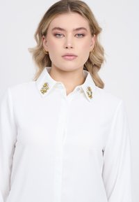 Camicia bianca con colletto decorato da accenti di gemme dorate e trasparenti. Tessuto liscio con maniche lunghe e chiusura a bottoni.