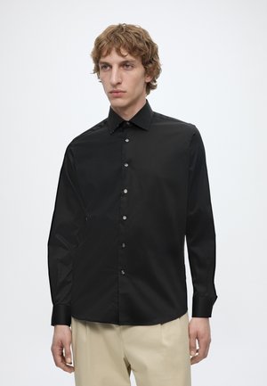 Junger Mann mit hellbraunen lockigen Haaren, der ein schwarzes Hemd mit Knöpfen und beige Hosen trägt und vor einem schlichten weißen Hintergrund steht.