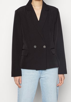 Femme portant un blazer noir croisé à deux boutons avec poches à rabat, associé à un jean bleu clair sur fond uni.