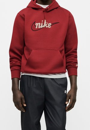 Mand iført en rød Nike hættetrøje med beige logo og sorte Nike bukser, stående mod en ensformet lys baggrund.