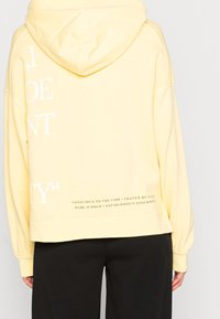 Felpa gialla con vestibilità rilassata, scollo con cappuccio e grande testo bianco sul retro. Presenta un orlo inferiore con dettagli stampati in nero.