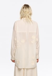 Camicia a maniche lunghe di colore crema in tessuto leggero, con ricami floreali sulla schiena, polsini con bottoni e una vestibilità comoda.