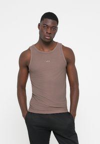 Brun ribbet tank top med rund hals, slim fit, med et tonalt logo på brystet. Sat sammen med sorte træningsbukser.