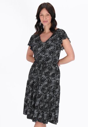 Femeie cu păr șaten mediu, purtând o rochie alb-negru cu flori și mâneci scurte, stând cu mâinile la spate.