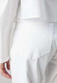 Personne portant un jean en denim blanc et une chemise blanche à manches longues, main détendue le long du corps sur un fond uni.