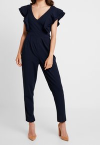 Námořnický overal s rozšířenými krátkými rukávy, hlubokým výstřihem do V a zúženými nohavicemi. Hladká tkanina s přiléhavým pasem. Kombinováno s nahými vysokými podpatky.