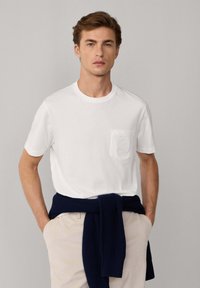 Camiseta de algodón blanca con cuello redondo y mangas cortas, que presenta un bolsillo con un diseño bordado. Combinada con pantalones de color crema y un suéter de punto azul marino atado a la cintura.