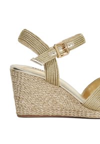Sandale compensée beige avec des détails de strap tressé et des accents métalliques. Fermeture par boucle sur la sangle de la cheville, avec une semelle compensée texturée.