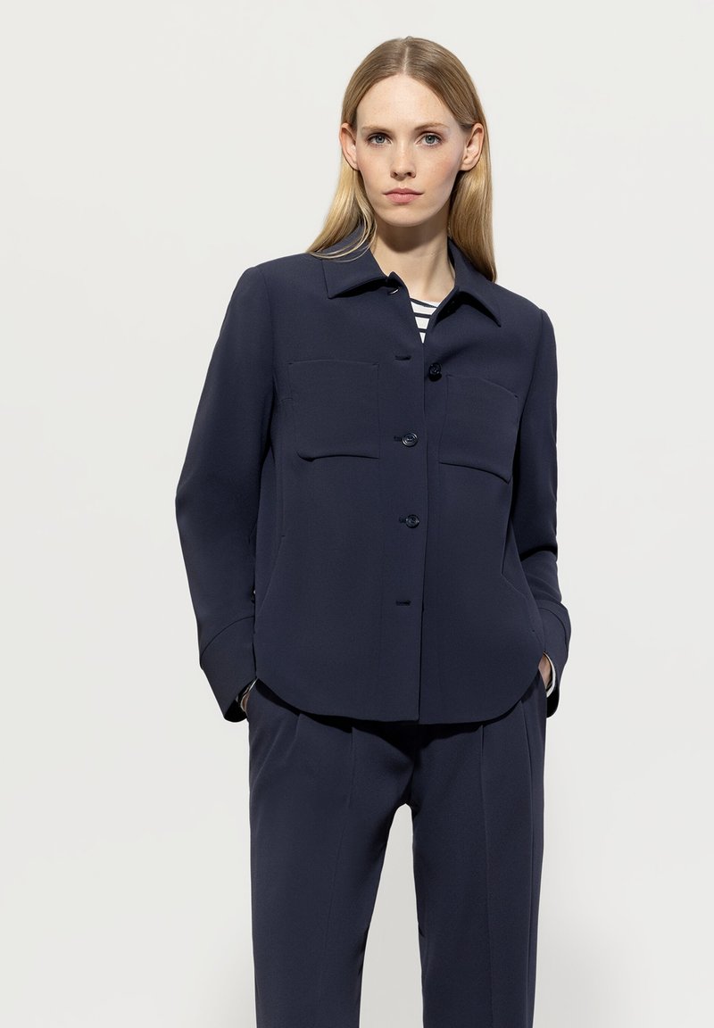 Navy blue button-up blouse with a collar, featuring two front pockets, loose fit, and structured shoulders; fabric appears smooth and lightweight.