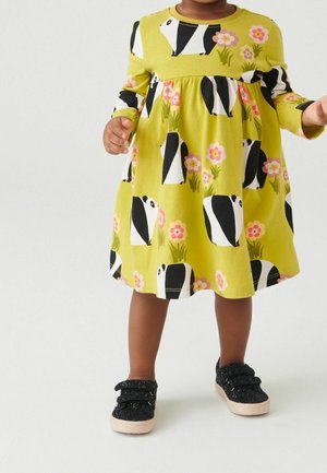 Kleid aus leuchtend grünem Stoff mit schwarz-weißen Dachsen und pinken Blumen. Lange Ärmel, geraffte Taille und lockere Passform. Kombiniert mit schwarzen Glitzerschuhen.