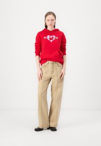 Červená bavlnená mikina s grafikou "NEW YORK", v kombinácii s béžovými širokými nohavicami. Texturovaná tkanina a uvoľnený strih. Čierne topánky dotvárajú outfit.