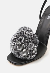 Svart høyhælte sandal med en stor, teksturert rosedesign laget av sølvfargede rhinestener på stroppen, hel tå og ankelstropper.