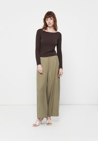 Top lung cu mânecă lungă de culoare maro, asortat cu pantaloni largi de un verde deschis. Modelul stă pe un fundal alb. Încălțămintea are un toc mic și un logo.