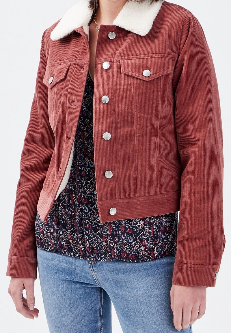 Femme portant une veste en velours côtelé couleur rouille avec col en polaire blanche, un chemisier à motif floral et un jean bleu clair, debout devant un fond uni.