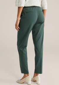 Sieviete valkā tumši zaļas sašaurinātas bikses un baltus tīkliņa stila kurpes ar biezām papēžām, stāvot pret vienkāršu neitrālu fonu.