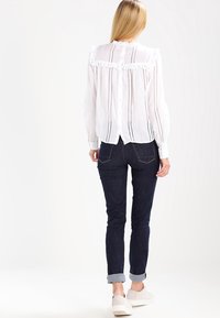 Vit skjorta med rynkad krage och vertikala öljett-dekoreringar, tillsammans med mörka denim jeans som är uppvikta vid ärmarna och vita skor.