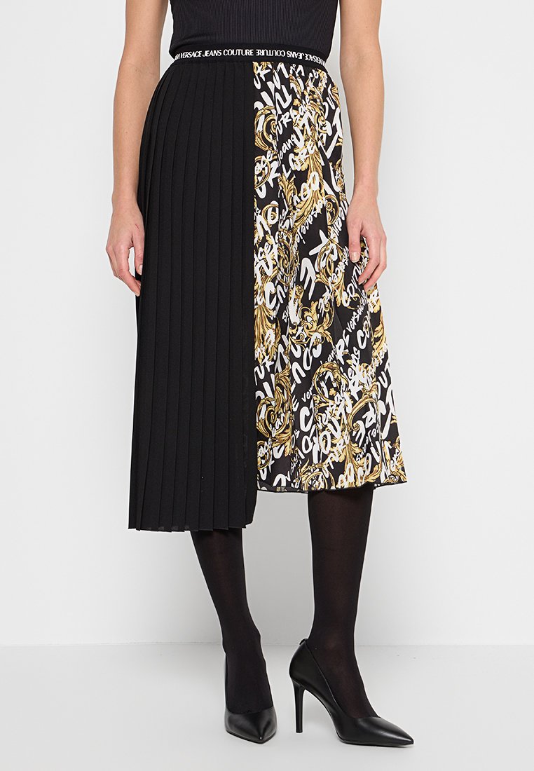 Schwarzer, plissierter Midirock, kombiniert mit einem schwarzen, weißen und goldenen gemusterten Panel, getragen mit schwarzen Strumpfhosen und schwarzen High Heels.