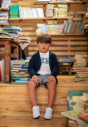 Niño sentado en unos escalones de madera rodeado de libros. Lleva un cárdigan azul marino, camisa blanca, pantalones cortos grises y zapatillas blancas.