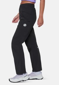 Γυναίκα που φοράει μαύρο εξωτερικό παντελόνι με φερμουάρ στην τσέπη του μηρού και λευκά παπούτσια πεζοπορίας με λογότυπο Mammut, στέκεται μπροστά σε μια μονόχρωμη επιφάνεια.