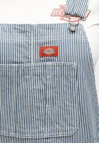Μπλε και άσπρες ριγέ φόρμες με μπροστινή τσέπη και κόκκινο έμβλημα Dickies, φορεμένες πάνω από ένα λευκό πουκάμισο με κόκκινο λογότυπο.