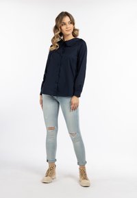 Marineblå skjorte med knapper og bølgende krave, lange ærmer og afslappet pasform, parret med lyseblå slidte jeans og beige ankelstøvler.
