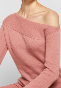 Pull en tricot rose doux à épaules dénudées, avec un encolure large, des manches longues et un motif texturé sur l'ensemble.