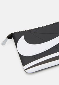 Čierno-biely Nike puzdro z hladkého syntetického materiálu, s výrazným bielym logom, zipsom a cik-cak švom na hornom okraji.